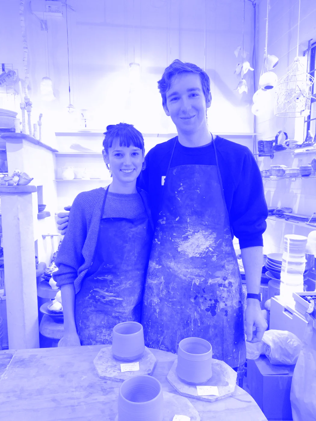 Jess and Patrick standing in a room full of pottery. In front of them in the 3 cups they made.